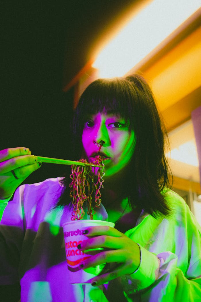 Portrait of a woman enjoying instant noodles under vibrant neon lights at night.